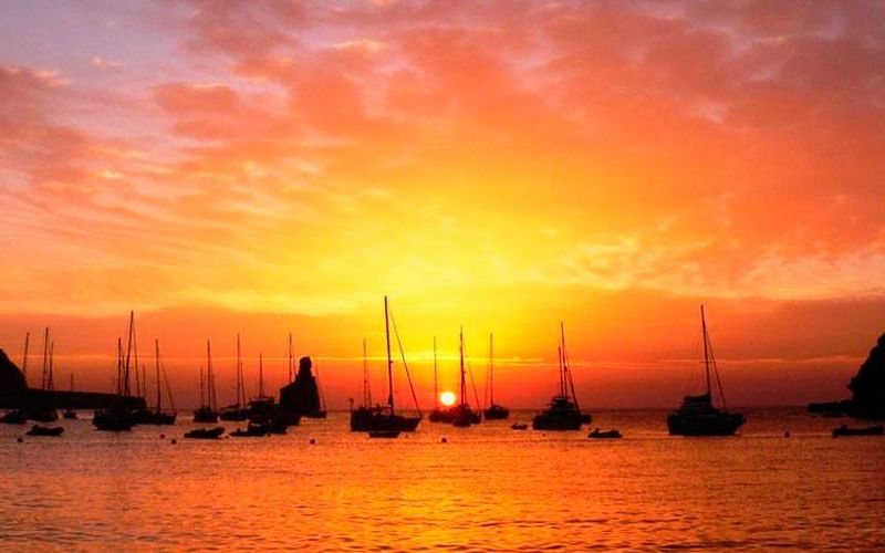 Atardecer en Cala Benirrás, con el cielo teñido de tonos naranjas, rojos y dorados sobre el mar. Varias embarcaciones fondeadas aparecen en silueta frente al horizonte, creando una escena cálida, mediterránea y muy vinculada a la magia del norte de Ibiza.